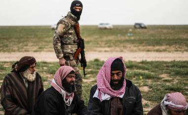 A fighter with the US-backed Syrian Democratic Forces (SDF) walks past men at a screening area for those who are evacuated from the Islamic State (IS) group's embattled holdout of Baghouz, during an operation to expel IS jihadists from the area, in the eastern Syrian province of Deir Ezzor, on February 26, 2019.