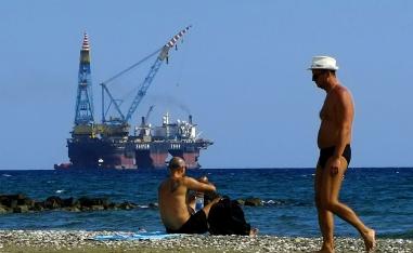 Men sit on a beach as a drilling platform is seen in the background near Larnaca